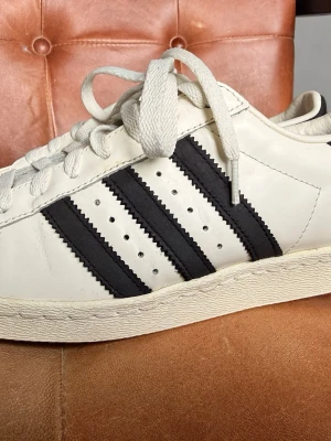 Adidas Superstar  - Klassiska Adidas Superstar sneakers i vitt läder med de ikoniska tre svarta ränderna på sidorna och snäckformad tå. Skorna har vita skosnören och en robust gummisula. Perfekta för en stilren och sportig look. Se bilderna för skicken.