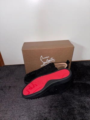 Svarta Louboutin sneakers med nitar - Säljer ett par svarta sneakers från Christian Louboutin med klassisk röd sula och svarta nitar på tån. Skorna är låga och har en lyxig känsla med mockaliknande material och svarta skosnören. Perfekta för dig som vill sticka ut med exklusiv design.