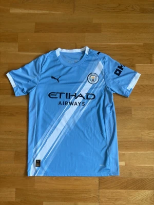 Manchester City De Bruyne matchtröja - Ljusblå Manchester City matchtröja från Puma med vita och mörkblå detaljer, klubbmärke på bröstet och sponsortryck. På ryggen står det 'De Bruyne' och nummer 17. Perfekt för fotbollsfans som vill visa sitt stöd på läktaren eller planen.