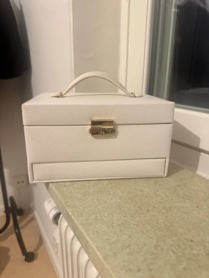 Vit smyckesbox med spegel - Stilren vit smyckesbox i läderimitation med guldfärgat lås och praktiskt handtag. Insidan har flera fack för ringar, örhängen och andra smycken samt en inbyggd spegel. Perfekt för att organisera dina accessoarer.