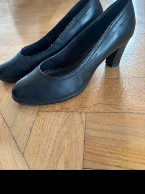 Svarta pumps från Tamaris i skinn - Klassiska svarta pumps från Tamaris i äkta skinn med rundad tå och medelhög klack. Enkla och stilrena med diskret söm längs kanten. Perfekta för dig som gillar tidlös och clean stil.