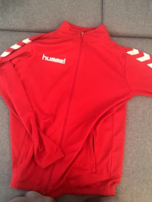 Röd träningsjacka från Hummel - Röd träningsjacka från Hummel med dragkedja framtill och vita detaljer på axlarna. Jackan har ståkrage, två fickor och är tillverkad i ett mjukt och sportigt material. Perfekt för träning eller uppvärmning. Passar till sommaren som kommer. Dragkedjan är dock lite konstig, men den funkar tillslut. 