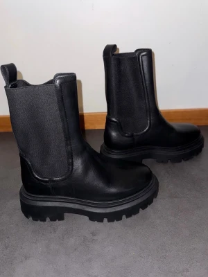 Svarta chunky Chelsea boots - Säljer ett par svarta Chelsea boots med grov sula och högt skaft. Skorna har elastiska paneler på sidorna och är tillverkade i slätt läder. Perfekta för dig som gillar en stilren men edgy look.