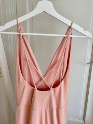Vacker lång ljusrosa klänning med smala band och öppen rygg - En fantastiskt fin långklänning från Bik Bok i en mjuk blush-rosa färg. Klänningen har V-ringning fram, öppen rygg med vackert korslagda smala band och en flödande kjol – en enkel och tidlös siluett som passar lika bra på ett bröllop som på en sommarkväll ute.  Klänningen är i mycket fint skick och har endast använts en kväll. Den är i storlek S, men är stretchig i materialet (jag har normalt sett M). 