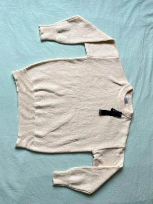 Krämvit stickad tröja Stone Island - Snygg krämvit stickad tröja från Stone Island med rund halsringning och ribbade muddar. Tröjan har en klassisk och enkel design som passar perfekt till många olika outfits. Materialet ser ut att vara mjukt och skönt, perfekt för kyligare dagar.