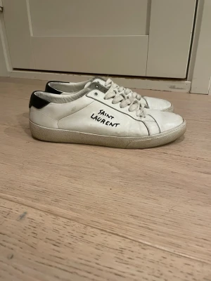 Vita Saint Laurent sneakers - Snygga vita sneakers från Saint Laurent med svart detalj på hälen och handskriven logga på sidan. Klassisk låg modell med vita snören och gummisula. Perfekta för dig som gillar stilrena och trendiga skor med exklusiv känsla.