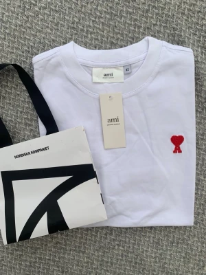Vit t-shirt från Ami med rött hjärta - Vit t-shirt från Ami Alexandre Mattiussi med ett broderat rött hjärta på bröstet. Klassisk rund halsringning och korta ärmar. Tillverkad i mjuk bomull för en skön känsla och stilren look.