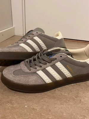 Adidas Gazelle grå sneakers - Grå Adidas Gazelle sneakers med klassiska vita ränder i textil och gummisula. Ovandelen är i mocka och skon har en låg profil. Guldtext med Gazelle-logga på sidan och blå etikett på plösen. Perfekta för en stilren och sportig look.