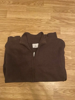 Brun ziptröja från Bläck - Mörkbrun stickad ziptröja från Bläck med hög krage och hel dragkedja framtill. Tröjan har ribbade muddar vid hals och ärmslut, perfekt för lager-på-lager. Enkel och stilren design som passar till det mesta.