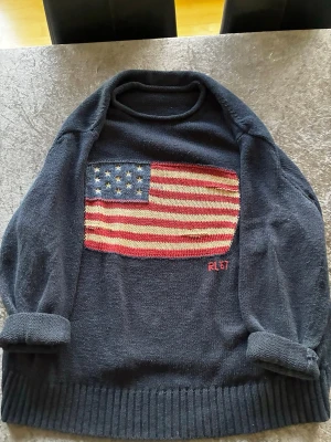 Mörkblå stickad tröja Polo Ralph Lauren - Mörkblå stickad tröja från Polo Ralph Lauren med stor amerikansk flagga på bröstet och RL67-brodyr. Tillverkad i en mix av linne och bomull, med ribbstickade muddar och avslappnad passform. Perfekt för dig som gillar klassisk och cool stil.