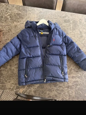 Blå pufferjacka från Polo Ralph Lauren - Snygg blå pufferjacka från Polo Ralph Lauren med huva och två dragkedjeförsedda fickor. Jackan har broderad röd logga på bröstet och quiltad design. Perfekt för kalla vinterdagar och riktigt skön att bära. Storlek 10-12 kille men passar mer åt de större hållet. Nästan aldrig använd 