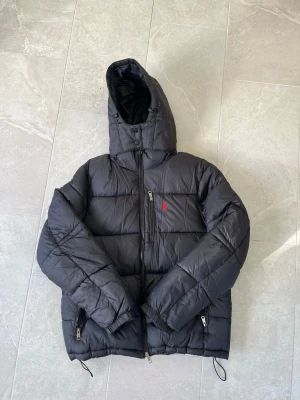 Svart pufferjacka från Polo Ralph Lauren - Svart pufferjacka från Polo Ralph Lauren med röd broderad logga på bröstet. Jackan har huva, dragkedjor på fickor och justerbara snören i nederkant. Perfekt för kalla vinterdagar och riktigt skön att bära. Klassisk och stilren design.