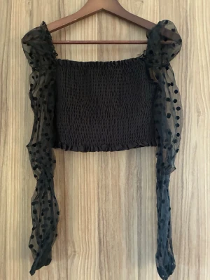 Svart mesh topp med puffärmar - Trendig svart topp med smockad kropp och långa puffärmar i transparent mesh. Ärmarna har ett prickigt mönster som ger en cool look. Perfekt att styla med jeans eller kjol för en snygg outfit.