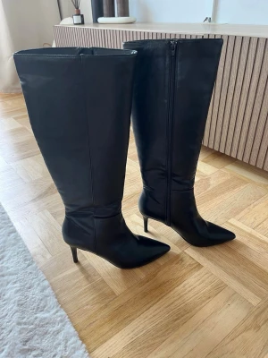 Svarta höga stövlar med klack - Snygga svarta stövlar med spetsig tå och smal klack. Stövlarna är knähöga och har dragkedja på insidan för enkel påtagning. Tillverkade i skinnliknande material med stilren design, perfekta för att lyfta vilken outfit som helst.