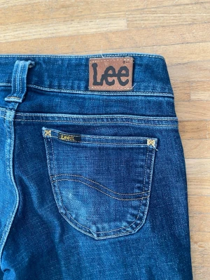 Blå bootcut jeans från Lee - Säljer ett par klassiska blå bootcut jeans från Lee med snygga detaljer och Lee-logga på bakfickan. Jeansen har normal passform och är tillverkade i bomull, perfekta för en avslappnad och trendig look.