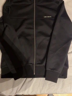 Svart zip-hoodie från Les Deux - Stilren svart hoodie från Les Deux med dragkedja framtill och diskret logga på bröstet. Tröjan har ribbade muddar vid ärmslut och nederkant. Perfekt för en avslappnad och trendig look.