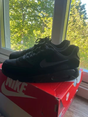  Nike Air Max 90 - Snygga helsvarta Nike Air Max sneakers med klassisk siluett och synlig Air-sula. Skorna har en låg profil, ovandel i textil och syntet samt diskret Nike-logga på sidan. Perfekta för dig som gillar stilrena och sportiga sneakers.