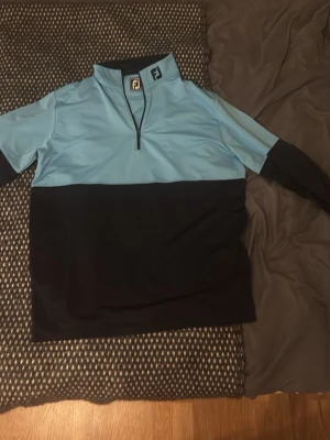 Blå och svart golftröja från FootJoy - Snygg långärmad golftröja från FootJoy i ljusblått och svart. Tröjan har halv dragkedja framtill och hög krage, perfekt för kyliga dagar på golfbanan. Tillverkad i ett stretchigt funktionsmaterial som ger bra rörelsefrihet.