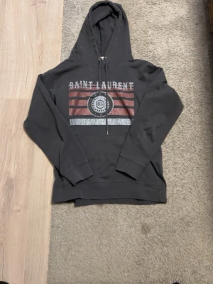Svart Saint Laurent hoodie med tryck - Säljer en svart hoodie från Saint Laurent med huva och dragsnören. På bröstet finns ett stort tryck med röda och vita ränder samt Saint Laurent-logga. Hoodien har en klassisk känguruficka och ribbade muddar vid ärmar och nederkant. Perfekt för en chill och stilren look.