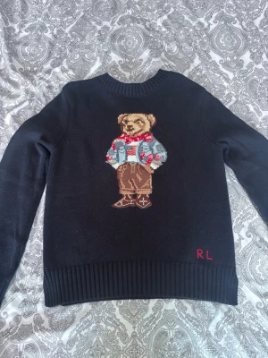 Svart stickad tröja med björn från RL - Svart stickad tröja från Ralph Lauren med ikonisk björn på bröstet, klädd i skjorta, röd scarf och bruna byxor. Tröjan har rund halsringning, ribbade muddar och RL-broderi nertill. Perfekt för dig som gillar klassiska och lekfulla detaljer.