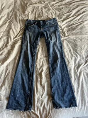 Supersnygga lågmidjade jeans - Säljer ett par blå bootcut jeans med rå, fransig kant längst ner. Jeansen har klassiska fickor fram och bak med snygga broderier på bakfickorna. Materialet är denim och passformen är utsvängd från knäna och ner.