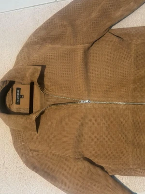 Brun skinnjacka från J Lindeberg  - Snygg brun skinnjacka från J Lindberg  med rak passform och klassisk krage. Jackan har hel dragkedja framtill och är tillverkad i perforerat skinn som ger en cool och modern look. Perfekt för dig som vill ha en stilren jacka med lite edge.