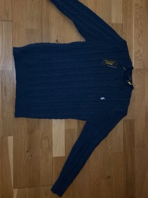Mörkblå kabelstickad tröja Polo Ralph Lauren - Mörkblå kabelstickad tröja från Polo Ralph Lauren med klassisk rund halsringning och broderad logga på bröstet. Tröjan har långa ärmar och ribbade muddar, perfekt för en stilren och tidlös look. Helt ny med lappen kvar aldrig använd. Pris kan diskuteras!