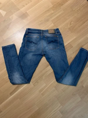 Blå slim fit jeans från Nudie - Säljer ett par klassiska blå slim fit jeans från Nudie med snygga slitningar och femficksmodell. Storlek: 30/32
