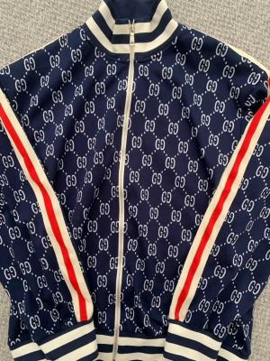 Blå zip-hoodie från Gucci med GG-mönster - Snygg blå zip-hoodie från Gucci med vitt GG-mönster över hela plagget. Ränder i vitt och rött längs ärmarna samt ribbade muddar och krage i blått och vitt. Tillverkad i ett mjukt och bekvämt material, perfekt för en trendig streetstyle-look.