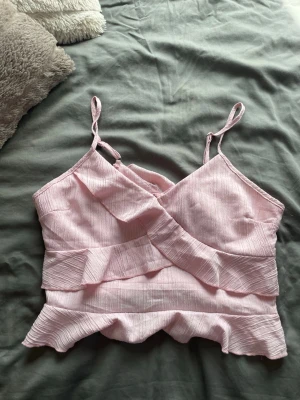 Ljusrosa topp med volanger - Supersöt ljusrosa topp med omlott-design och volangdetaljer. Tunt, ribbat material och smala axelband ger en feminin och somrig känsla. Perfekt att matcha med kjol eller jeans🥰