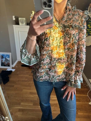 Blommig volangblus med smockdetaljer - Blus med färgglatt blommönster i grönt, orange och rosa toner. Volangdetaljer vid axlar och smock upptill, samt vida långärmar med dekorativ kant. Luftig och lätt i materialet, perfekt för en trendig och avslappnad look.
