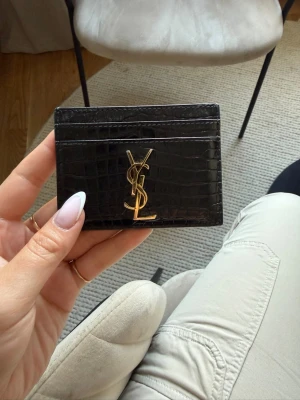 Svart YSL korthållare i läder - Snygg svart korthållare från Saint Laurent i croco-präglat läder med guldigt YSL-logo framtill. Smidig och stilren design med flera kortfack, perfekt för att hålla koll på dina viktigaste kort.