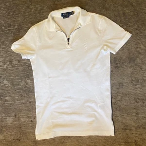 Vit polotopp från Polo Ralph Lauren - Vit polotopp från Polo Ralph Lauren med klassisk krage och half zip-dragkedja framtill. Broderad logga på bröstet och korta ärmar. Perfekt för en clean och stilren look.