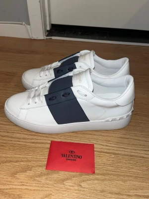 Valentino Garavani vit/mörkblå skor - Stilrena vita sneakers från Valentino Garavani med bred mörkblå rem över vristen och ikoniska nitar på hälen. Skorna är i läder och har en chunky sula som ger en modern vibe. Perfekta för dig som vill sticka ut med exklusiva detaljer.