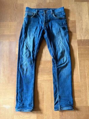 Blå jeans från Nudie Jeans - Säljer ett par blå Grim Tim jeans från Nudie i storlek 30/30. Jeansen har snygga slitningar framtill och bakfickor med Nudie Jeans signaturbrodyr. Knappt använda så är i toppskick!