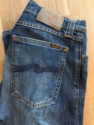 Blå jeans från Nudie Jeans/narrow boot blue nightrider - Snygga blå jeans från Nudie Jeans med klassisk femficksdesign och tydliga orange sömmar. Jeansen har en mörkblå tvätt och vågig brodyr på bakfickan. Tillverkade i denim av bomull för en skön och avslappnad look.