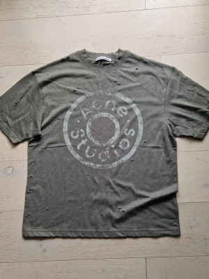 Grå Acne Studios t-shirt med tryck - Cool grå t-shirt från Acne Studios med stort, slitet logotryck framtill. Oversized passform och korta ärmar. T-shirten har distressed detaljer för en edgy look och är tillverkad i mjuk bomull.