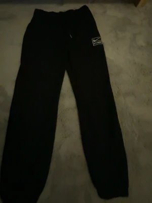 Svarta Stussy x Nike mjukisbyxor - Säljer ett par svarta mjukisbyxor från Stussy x Nike med broderad logga på benet. Byxorna har resår i midjan och vid bensluten för en skön och avslappnad passform. Perfekta för chill eller streetwear-stil.