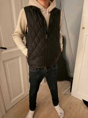 Svart quiltad dunväst med dragkedja - Snygg svart dunväst med quiltat mönster och hög krage. Västen har tvåvägsdragkedja framtill och två sidofickor. Perfekt att slänga över en hoodie för en chill look. Materialet är lätt och ger bra värme utan att kännas klumpig.