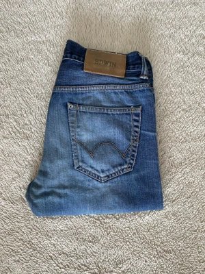 Mörkblå jeans från Edwin - Säljer ett par mörkblå jeans från Edwin med snyggt slitna detaljer och klassisk femficksmodell. Jeansen har raka ben och är tillverkade i kraftig denim med en cool tvätt som ger en avslappnad look. Jeansen har ett litet hål/slitning vid skrevet.