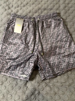 Grå Fendi badbyxor med logomönster - Snygga grå badbyxor från Fendi med ikoniskt FF-logomönster över hela tyget. De har elastisk midja med svarta dragsnören och innerfoder i mesh. Perfekta för stranden eller poolen om du vill sticka ut med lyxig stil. PASSAR M. HELT NY, OANVÄND.