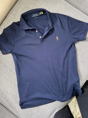 Marinblå pikétröja från Polo Ralph Lauren - Klassisk marinblå pikétröja från Polo Ralph Lauren med broderad logga på bröstet. Tröjan har krage, korta ärmar och knäppning med tre knappar framtill. Perfekt till jeans eller shorts för en stilren look.