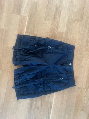 Svarta shorts med snörning -  Svarta shorts med coola detaljer som snörning och stora fickor både fram och bak. De har en avslappnad passform och är perfekta för varma dagar. Stängs med knapp och dragkedja framtill.