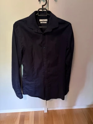 Mörkblå skjorta från Jack and Jones - Stilren mörkblå skjorta från Jack and Jones med klassisk krage och knäppning framtill. Skjortan har lång ärm och är tillverkad i mjuk bomull som känns skön mot huden. Perfekt för dig som gillar en clean och enkel look.