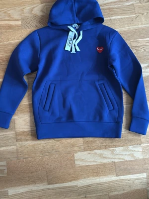 Blå hoodie från Comme des Garçons Play - Snygg blå hoodie från Comme des Garçons Play med det ikoniska röda hjärtat med ögon broderat på bröstet. Hoodien har huva med dragsko och en stor magficka. Perfekt för en chill och trendig look.