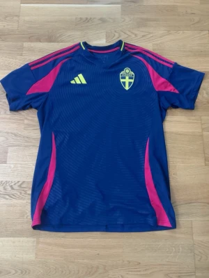 Blå Sverige Adidas matchtröja - Säljer en blå matchtröja från Adidas med Sveriges landslagsemblem på bröstet. Tröjan har gula och rosa detaljer, korta ärmar och texten 'Sverige' i nacken. Perfekt för fotboll eller träning.