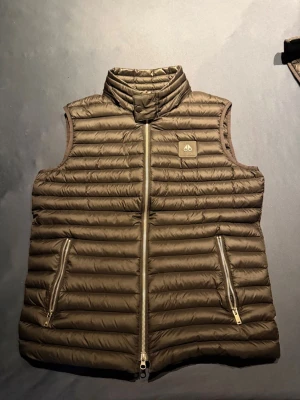 Svart dunväst från Moose Knuckles - NYPRIS - 5000kr.                                  Snygg brun dunväst från Moose Knuckles med quiltad design och hög krage. Västen har två dragkedjeförsedda fickor och logotyp på bröstet. Tillverkad i lätt och vattenavvisande material med dunfyllning för extra värme. Perfekt för lager-på-lager-stil.