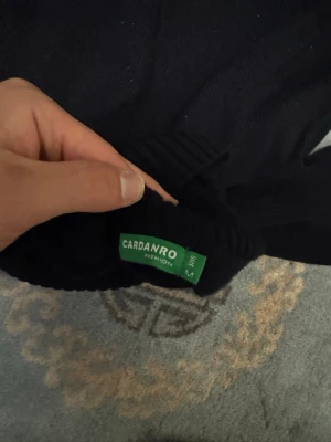 Svart polotröja med blå ränder Cardanro - Snygg svart stickad polotröja från Cardanro med tre ljusblå horisontella ränder över bröstet. Tröjan har lång ärm, ribbstickade muddar och hög polokrage. Perfekt för dig som gillar en stilren och enkel look.
