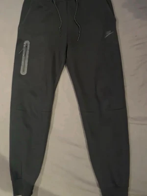 Svarta Nike tech byxor - Svarta Nike tech byxor. Byxorna har en snygg dragkedjeficka på ena benet och Nike-logga på låret. Perfekta för chill eller träning. De är helt oanvända!