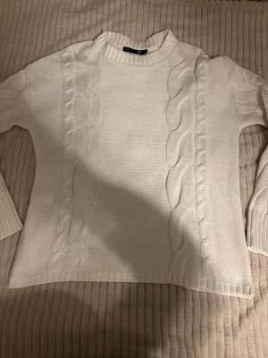 Vit kabelstickad tröja  - Stickad vit tröja från lager 157 med rund halsringning och ribbade muddar. Tröjan har ett fint mönster framtill och baktill samt långa ärmar. Perfekt för lager på lager och enkel att matcha med jeans eller kjol.